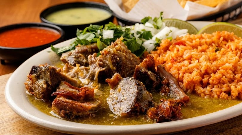 Braised pork in green sauce with red rice, cilantro, onions, lime, and two bowls of salsa.